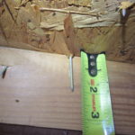 Close-up image of a nail embedded in wooden decking, with a tape measure showing the nail's length.